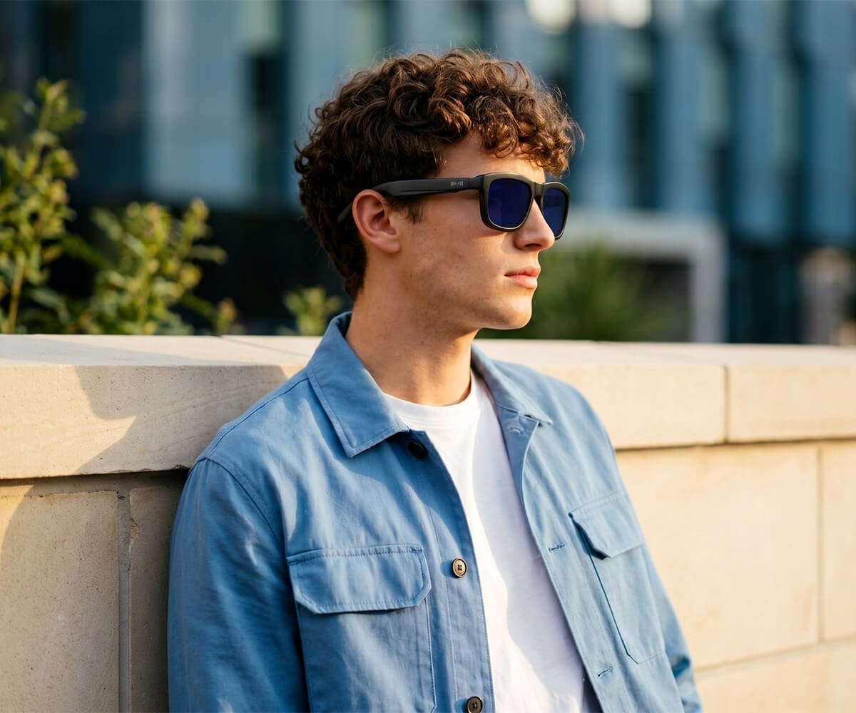 A man with curly brown hair wearing black sunglasses with dark blue lenses, a light blue denim shirt, and a white t-shirt, leaning against a beige stone wall in an outdoor urban setting.