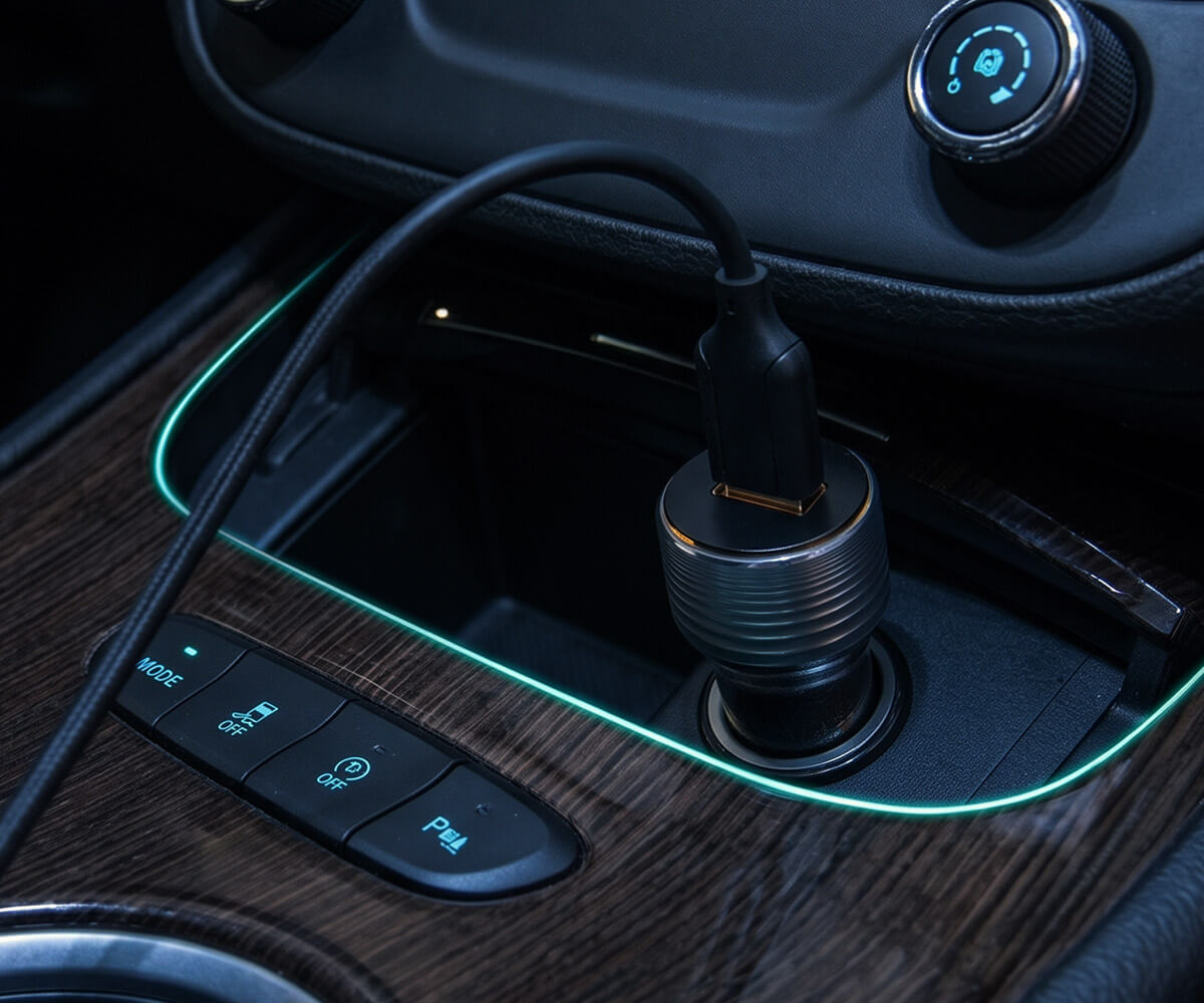 A black car adapter plug with a metallic silver threaded base, shown inserted into a car's 12V socket. The socket is set in a dark wood console with ambient green lighting, and a black charging cable is plugged into the adapter.