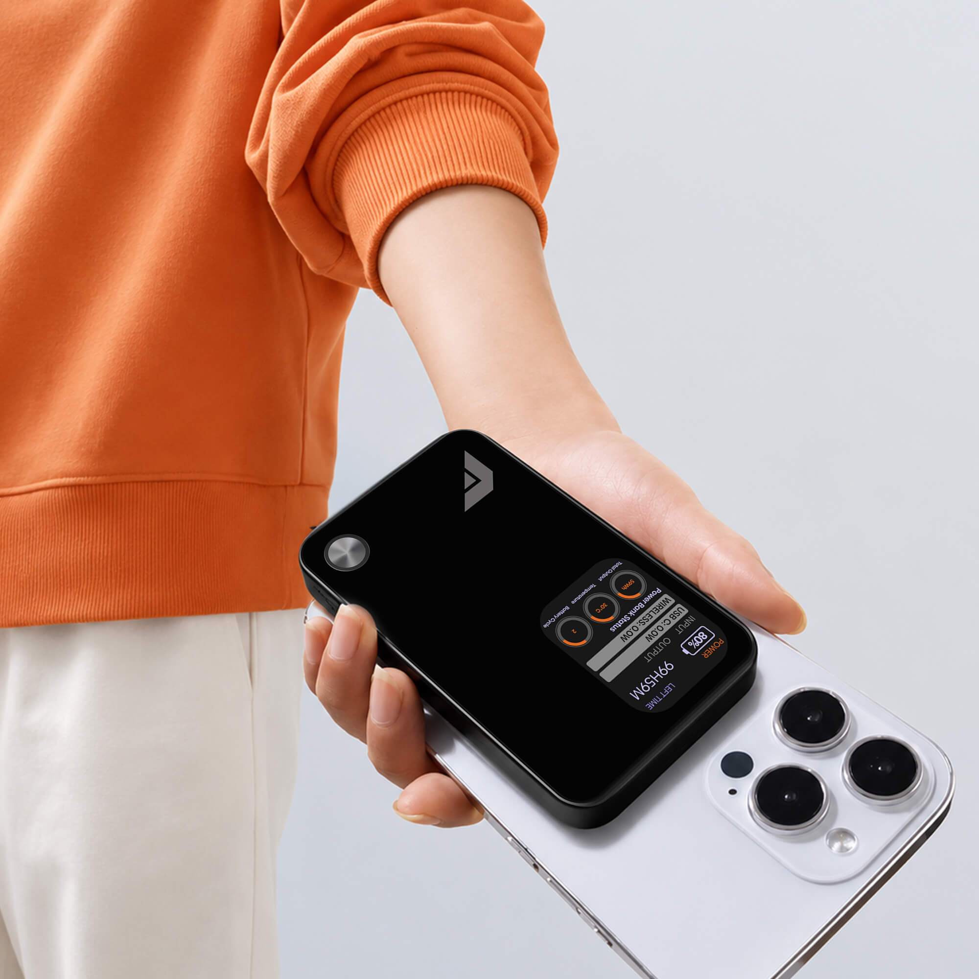 A person in an orange sweatshirt holds a black rectangular phone accessory attached to a white smartphone against a light gray studio background.