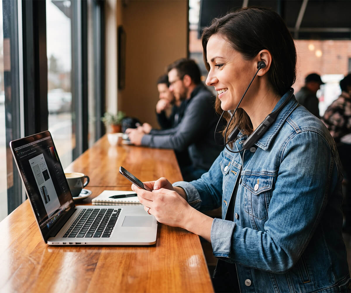 A woman in a blue denim jacket sits at a wooden table in a cafe, wearing a faster neckband and looking at her phone next to an open laptop.