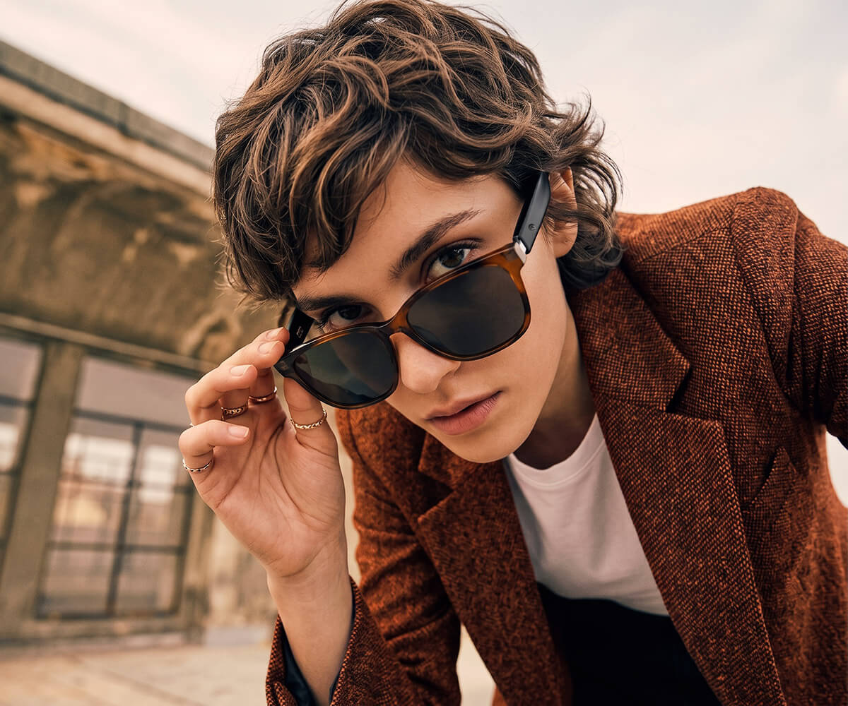 A close-up lifestyle shot of a person with wavy brown hair wearing leapard sunglasses and a brown tweed blazer over a white t-shirt, holding the sunglasses in front of their face against an outdoor architectural background.