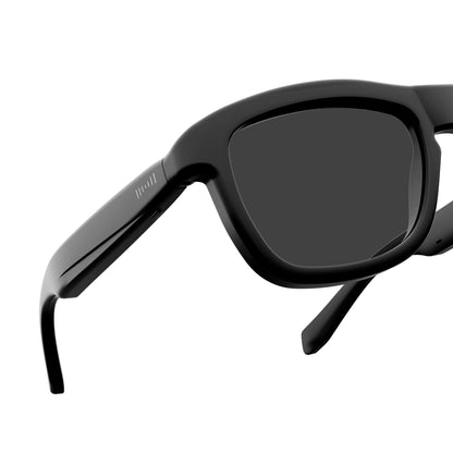 Close-up of black sunglasses with dark lenses and a visible brand mark on the temple arm, set against a plain white background.