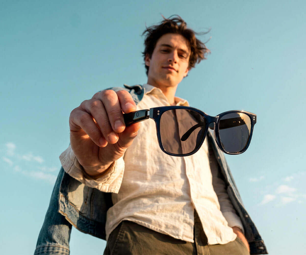 A man outdoors holding up a pair of black-framed sunglasses toward the camera against a blue sky background.