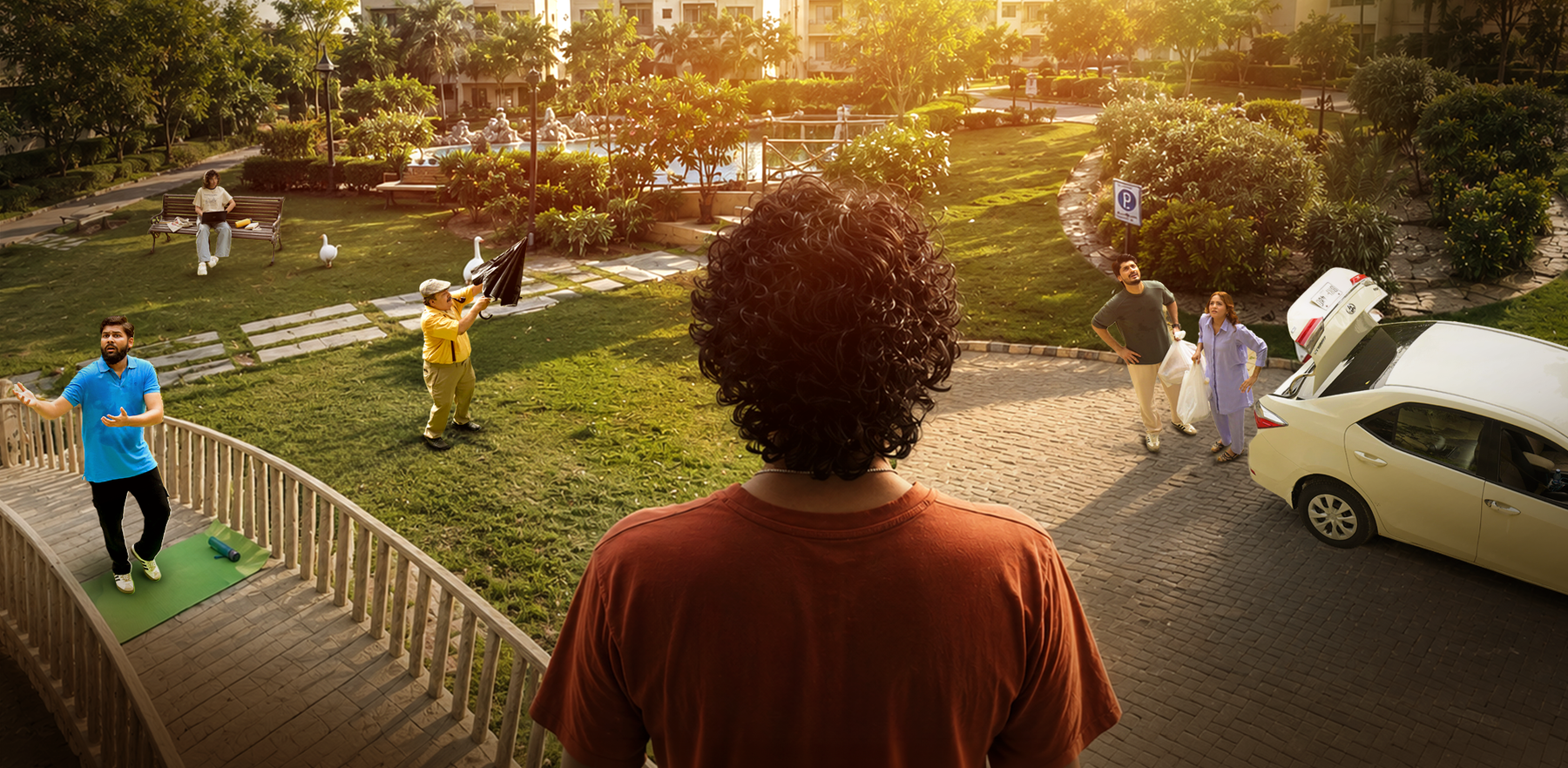 A man with curly dark hair seen from behind, wearing a rust-orange t-shirt, standing in a sunny outdoor garden courtyard. In the foreground, a woman in a blue shirt does yoga on a green mat on a wooden deck.