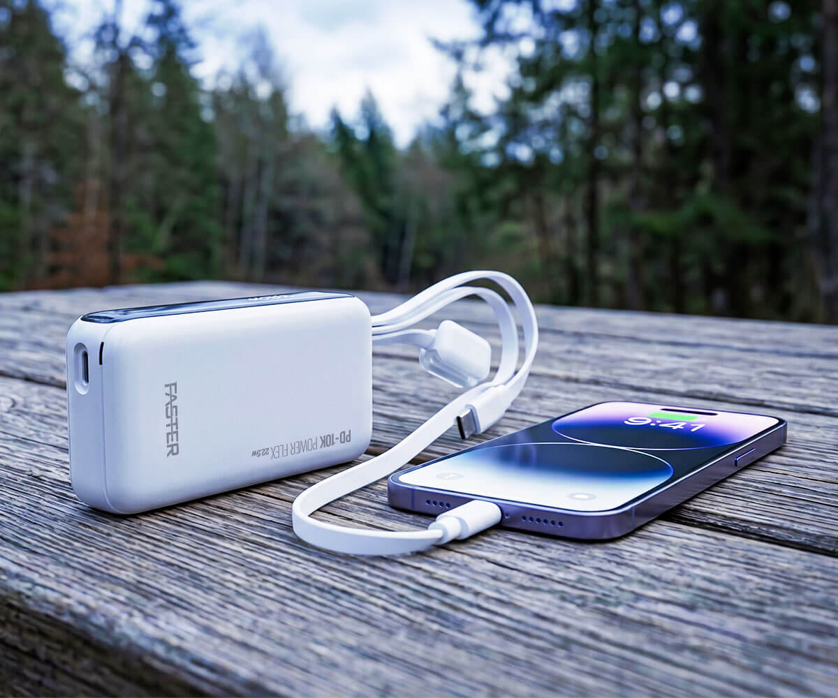 A white Master portable power bank connected to a smartphone via a white charging cable, placed on a weathered wooden table with a blurred forest background.